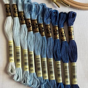 Op de afbeelding: Een verzameling blauwe borduurgaren in diverse tinten, van licht tot donker, op een witte ondergrond. Een houten borduurring is zichtbaar. De garens zijn voorzien van nummers.