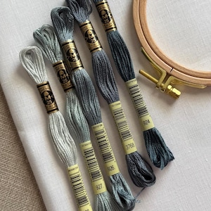 May include: An assortment of six skeins of embroidery floss in shades of gray and blue, with a wooden embroidery hoop. The floss is labeled with numbers and brand information. The hoop is approximately 6 inches in diameter.