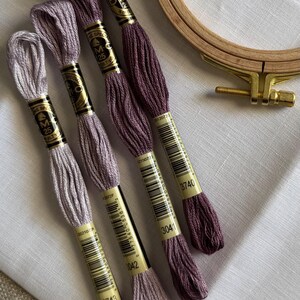 May include: An assortment of embroidery floss in shades of purple and lavender, with a wooden embroidery hoop. The floss is labeled with numbers and brand information. The items are arranged on a white fabric.
