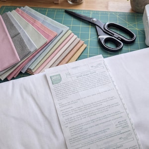 Könnte beinhalten: Ein Stapel gefalteter Stoffquadrate in Rosa-, Grau- und Blautönen, eine schwarze Nähschere und ein weißer Bogen Pellon SF101 Shape-Flex Bügeleinlage mit gedruckter Anleitung, alles auf einer grünen Schneidematte platziert.