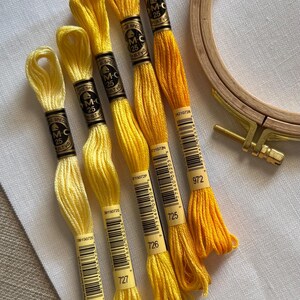 May include: A collection of yellow embroidery floss skeins in varying shades, from pale to vibrant, with a wooden embroidery hoop. The floss is labeled with numbers and brand information, ideal for needlecraft projects.