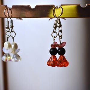 Puede incluir: Tres pares de pendientes colgando de un expositor. Los pendientes están hechos de cuentas y tienen diferentes colores y diseños. Los pendientes de la izquierda son blancos e iridiscentes, los pendientes del centro son rojos y negros, y los pendientes de la derecha son rosas y negros.