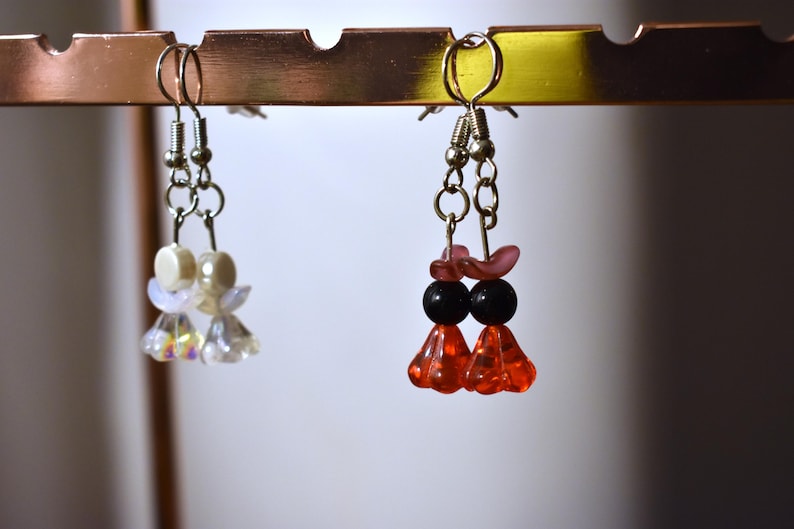 May include: Three pairs of earrings hanging on a display rack. The earrings are made of beads and have different colors and designs. The earrings on the left are white and iridescent, the earrings in the middle are red and black, and the earrings on the right are pink and black.