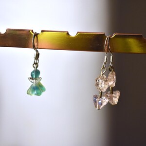 May include: Two pairs of earrings hanging on a gold metal rack. The earrings on the left are green and blue with a flower design. The earrings on the right are clear and pink with a crystal design.