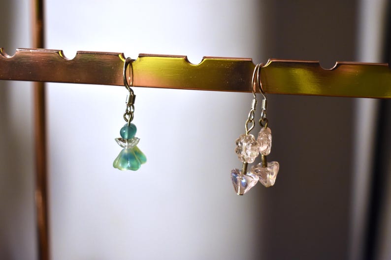 May include: Two pairs of earrings hanging on a gold metal rack. The earrings on the left are green and blue with a flower design. The earrings on the right are clear and pink with a crystal design.