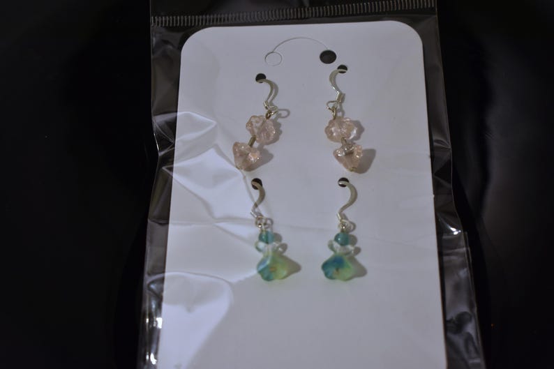 May include: A pair of earrings with pink and blue glass beads. The earrings are on a white card with a clear plastic backing.