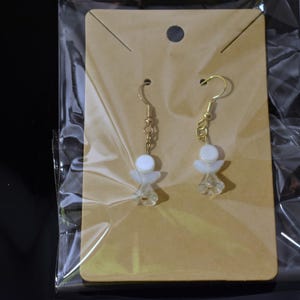 May include: A pair of gold dangle earrings with white angel-shaped charms. The earrings are displayed on a brown card with a clear plastic backing.