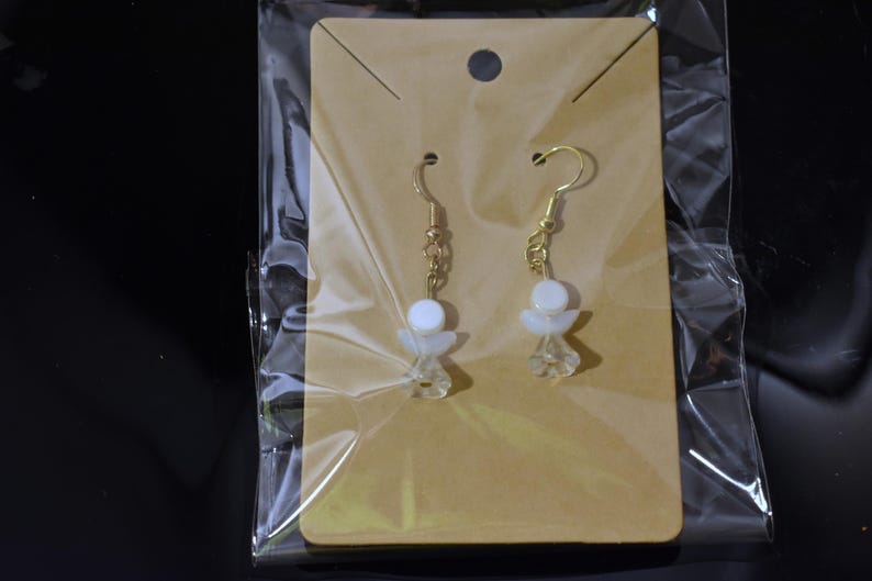 May include: A pair of gold dangle earrings with white angel-shaped charms. The earrings are displayed on a brown card with a clear plastic backing.