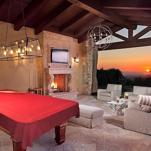 May include: A luxurious outdoor living space featuring a red pool table, a fireplace with a lit fire, and comfortable seating. The space is illuminated by hanging lights and offers a scenic view of a sunset.