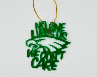 Op de afbeelding: Groen ornament met de tekst "NO ONE WE DON'T CARE" en een witte adelaarskop. Het ornament heeft een gouden touwtje om op te hangen. Het ontwerp is een gedurfde, grafische stijl.