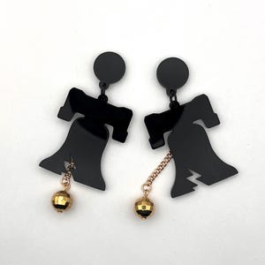 May include: A pair of black acrylic earrings shaped like the Liberty Bell with gold-toned dangling balls.