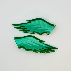 Könnte beinhalten: Zwei grüne, flügelförmige Accessoires mit glänzendem Finish. Die Flügel haben ein geschichtetes Design mit einem helleren grünen Innenraum und einer dunkleren grünen Kontur. Die Accessoires sind wahrscheinlich für dekorative Zwecke oder als Teil eines Kostüms.