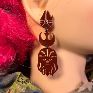 Puede incluir: Pendientes colgantes de acrílico marrón con diseños de Star Wars. Los pendientes muestran el Halcón Milenario, el símbolo de la Alianza Rebelde y la cara de Chewbacca. Están conectados por pequeñas cadenas negras.