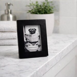 May include: A black rectangular picture frame with a black and white image of a pug dog sitting on a toilet and reading a newspaper. The frame is on a white marble countertop.