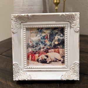 May include: A square, white, distressed picture frame with ornate corner detailing. The framed image depicts a pug dog lying near a Christmas tree and wrapped gifts. The frame is sitting on a dark wooden surface.