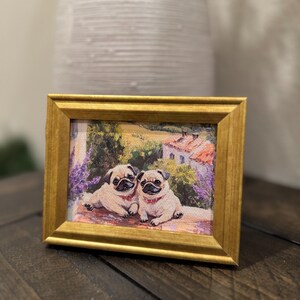 May include: A framed print featuring two pug dogs with red collars, set against a backdrop of a European landscape with a house and lavender fields. The frame is gold-colored, and the artwork is displayed on a dark wooden surface.