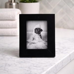 May include: A black framed photograph of a pug dog in a bathtub covered in bubbles. The black and white image is set on a white marble countertop, with a stack of white towels and a small plant in the background.