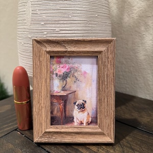 May include: A small framed print featuring a pug dog sitting in front of a table with a vase of flowers. The frame is a light brown color. A lipstick tube is next to the frame.