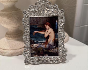 May include: Ornate silver-toned picture frame with a painting of a mermaid with long red hair. The frame has a detailed, decorative design with small embellishments. The painting depicts a mermaid near the ocean.
