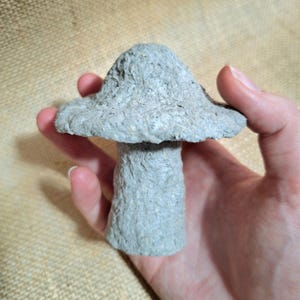 May include: A handmade, light gray mushroom sculpture with a textured surface. The mushroom has a wide, flat cap and a cylindrical stem. The sculpture is held in a hand.