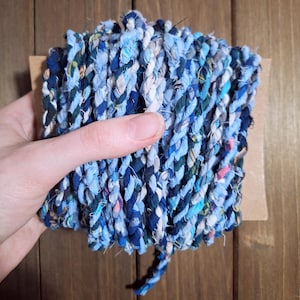 May include: A tightly coiled, braided rope made from various shades of blue fabric scraps. The rope is held against a brown cardboard backing, showcasing a mix of light and dark blues, with hints of other colors.
