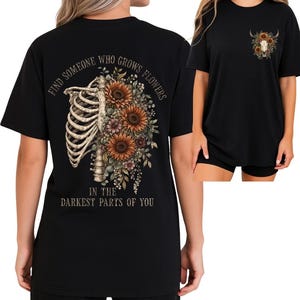 May include: Black t-shirt featuring a floral and anatomical design. The back of the shirt displays a rib cage with sunflowers and other flowers. Text on the back reads "FIND SOMEONE WHO GROWS FLOWERS IN THE DARKEST PARTS OF YOU". The front has a smaller design.