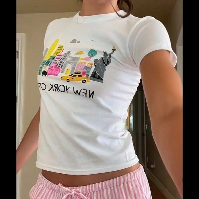 May include: White t-shirt with a colorful graphic of the New York City skyline, including the Statue of Liberty and a yellow taxi cab. The text "NEW YORK CITY" is printed below the graphic.