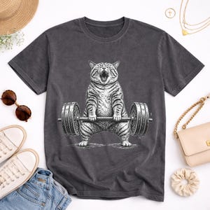 Könnte beinhalten: Dunkelgraues T-Shirt mit einer Schwarz-Weiß-Illustration einer Katze, die Gewichte hebt. Die Katze ist mit offenem Maul dargestellt, als würde sie brüllen. Die Langhantel hat auf jeder Seite zwei Gewichtsscheiben. Das T-Shirt hat einen Rundhalsausschnitt.
