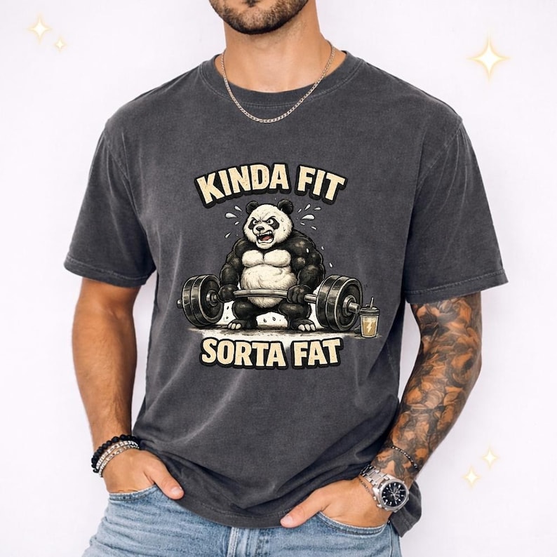 May include: Dark gray t-shirt featuring a cartoon panda lifting weights. The panda is black and white with an angry expression. The text "KINDA FIT" is above the panda, and "SORTA FAT" is below.