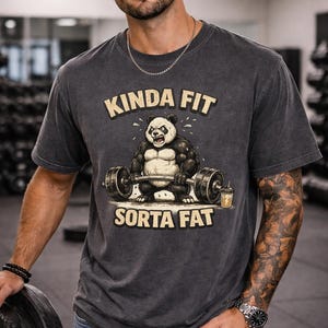 May include: Dark gray t-shirt with the text "KINDA FIT SORTA FAT" above a cartoon panda lifting weights. The panda is black and white with muscular arms. The shirt is worn by a person in a gym setting.