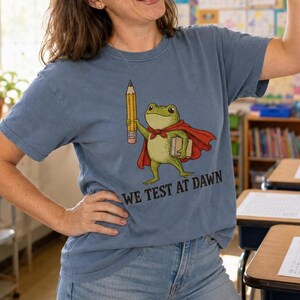 Puede incluir: Camiseta azul con un dibujo de una rana de dibujos animados con una capa roja, un lápiz y un libro. El texto "WE TEST AT DAWN" está impreso debajo. La camiseta es de material suave.