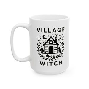 May include: White ceramic mug with the words "VILLAGE WITCH" in black, with a black illustration of a house, cat, mushrooms, and foliage. The mug has a large handle and is designed for hot beverages.