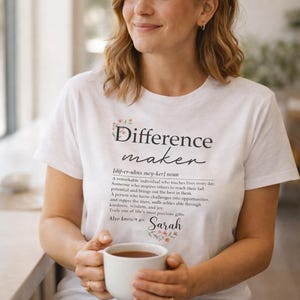 Op de afbeelding: Wit T-shirt met de tekst "Difference maker" en een definitie. Het shirt bevat ook de naam "Sarah". De persoon houdt een witte mok met een bruine vloeistof vast.