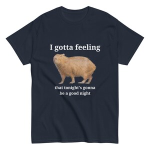 Pu&ograve; includere: Una t-shirt blu navy con un'illustrazione di capibara bianca e il testo "I gotta feeling that tonight's gonna be a good night".