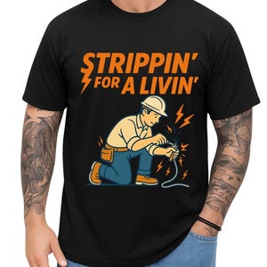 May include: Black t-shirt featuring the text "STRIPPIN' FOR A LIVIN'" in orange, with a lightning bolt graphic. The design includes an electrician in a hard hat, tan shirt, blue trousers, and orange boots.