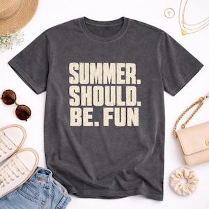 Könnte beinhalten: Ein anthrazitfarbenes Kurzarm-T-Shirt mit dem Text "SUMMER. SHOULD. BE. FUN", der in einer abgenutzten, cremeweißen Blockschrift mittig auf der Brust aufgedruckt ist. Das T-Shirt hat einen Rundhalsausschnitt und eine normale Passform.