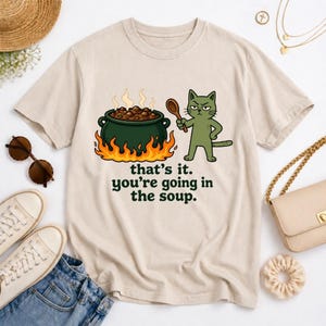 May include: Beige t-shirt featuring a cartoon cat holding a wooden spoon, standing next to a cauldron of soup over flames. The text "that's it. you're going in the soup." is printed below the cauldron. The shirt is a casual wear item.