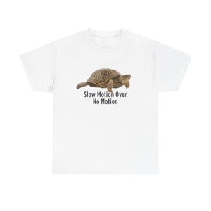 Op de afbeelding: Wit t-shirt met een gedetailleerde illustratie van een schildpad. De schildpad heeft een bruin schild en ledematen. De tekst "Slow Motion Over No Motion" staat eronder in donkergrijs.
