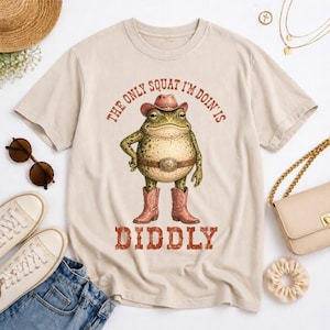 May include: Beige t-shirt featuring a cartoon frog wearing a cowboy hat, belt, and boots. The text "THE ONLY SQUAT I'M DOIN' IS DIDDLY" is printed above and below the frog illustration in a red, Western-style font. The shirt has short sleeves.