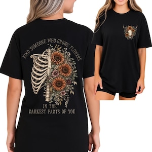May include: Black t-shirt featuring a floral design with sunflowers, a rib cage, and the text "FIND SOMEONE WHO GROWS FLOWERS IN THE DARKEST PARTS OF YOU." A smaller design of a skull with flowers is on the front.