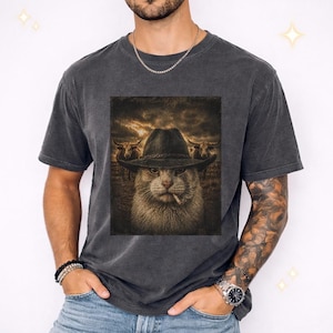 May include: Dark gray t-shirt featuring a graphic of a cat wearing a cowboy hat and smoking a cigarette, with two longhorn cattle in the background. The cat has a serious expression. The shirt is a vintage style.