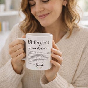 May include: White ceramic mug with the text "Difference maker" and a definition. The mug also includes the name "Sarah" and floral accents. The mug is held by a person.