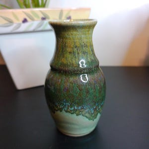 May include: A small, handcrafted ceramic vase with a unique, textured glaze. The vase features a gradient of green and brown hues, with a lighter green base. The design includes a distinct band around the middle. The vase is approximately 5 inches tall.