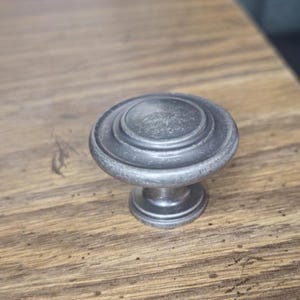 May include: A weathered, silver-toned cabinet knob with a circular design. The knob has a rounded top with concentric rings and a short, cylindrical base. The hardware is set against a wooden surface with a warm, brown tone.