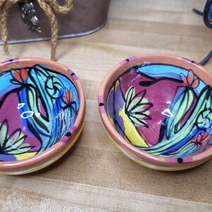 May include: Two small, hand-painted ceramic bowls. The interior features a vibrant design with purple, blue, yellow, and green floral motifs. The rims are a light orange color, and the bowls are set on a wooden surface.