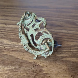 May include: An ornate, antique-style drawer pull with a distressed appearance. The metal hardware has a detailed, openwork design with a greenish-gold finish. Includes a screw for mounting. Suitable for vintage furniture restoration.