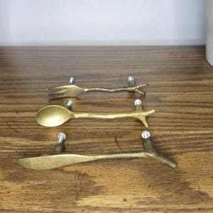 May include: Three brass-toned metal cabinet pulls shaped like miniature cutlery. The top pull resembles a fork, the middle a spoon, and the bottom a knife. Each piece features a textured, branch-like handle design with two silver-toned mounting screws.
