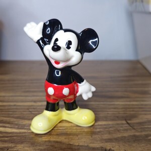 K&ouml;nnte beinhalten: Eine Keramikfigur von Mickey Mouse in einer klassischen Pose, mit schwarzen Ohren, einem schwarzen Torso, roten Shorts mit wei&szlig;en Kn&ouml;pfen und gelben Schuhen. Die Figur hat wei&szlig;e Handschuhe und ein l&auml;chelndes Gesicht. Die Figur ist etwa 10 cm gro&szlig;.
