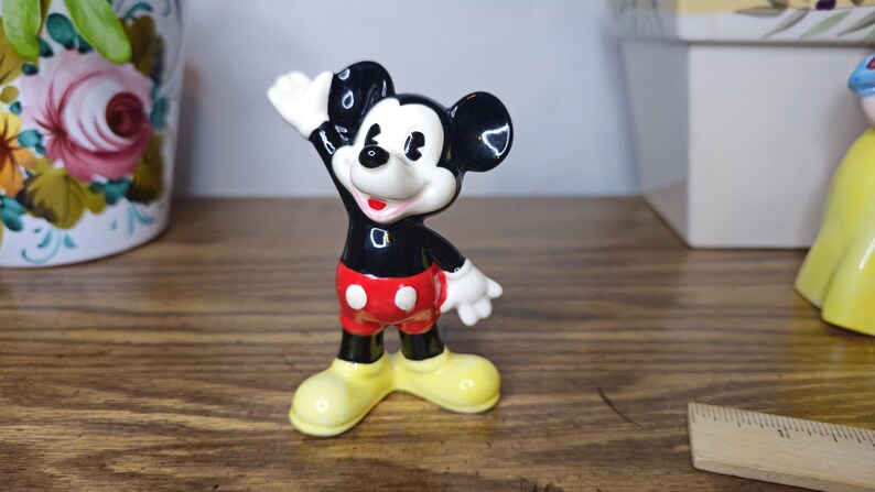 K&ouml;nnte beinhalten: Eine Keramikfigur von Mickey Mouse in einer klassischen Pose, mit schwarzen Ohren, einem schwarzen Torso, roten Shorts mit wei&szlig;en Kn&ouml;pfen und gelben Schuhen. Die Figur hat wei&szlig;e Handschuhe und ein l&auml;chelndes Gesicht. Die Figur ist etwa 10 cm gro&szlig;.