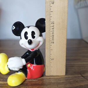 K&ouml;nnte beinhalten: Eine Keramikfigur von Mickey Mouse mit schwarzen Ohren, einem schwarzen K&ouml;rper, roten Shorts und gelben Schuhen. Die Figur ist etwa 7,6 cm hoch, mit wei&szlig;en Handschuhen und einem l&auml;chelnden Gesicht. Ein Holzlineal befindet sich daneben.
